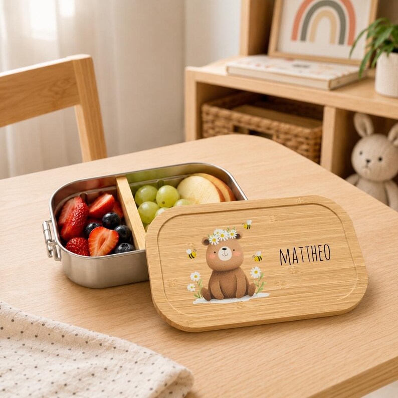May include: A rectangular stainless steel lunch box with a removable bamboo divider and a matching bamboo lid. The lid features a printed illustration of a brown bear wearing a daisy crown, surrounded by bees, and the name MATTHEO in black text.