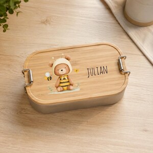 May include: A rectangular lunch box with a light brown bamboo lid featuring a printed illustration of a brown teddy bear wearing a yellow and black striped bee costume, accompanied by a small bee and the name JULIAN printed in black text.