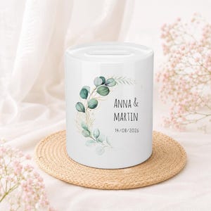 May include: A white ceramic money box featuring a cylindrical shape with a coin slot on top. The front displays a printed design of green eucalyptus leaves and gold line art branches, with the text ANNA & MARTIN and the date 14/08/2026.