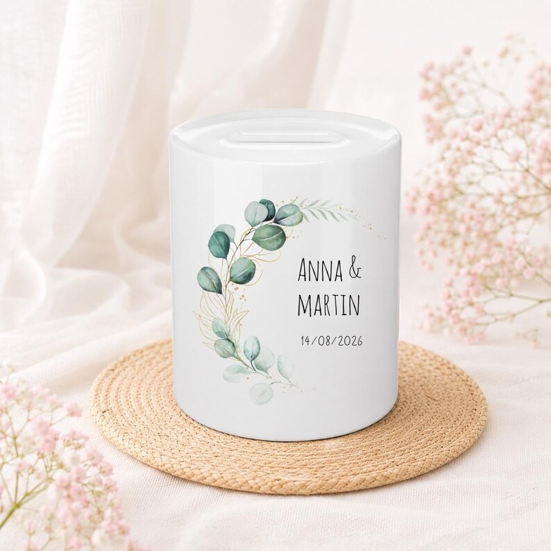 May include: A white ceramic money box featuring a cylindrical shape with a coin slot on top. The front displays a printed design of green eucalyptus leaves and gold line art branches, with the text ANNA & MARTIN and the date 14/08/2026.
