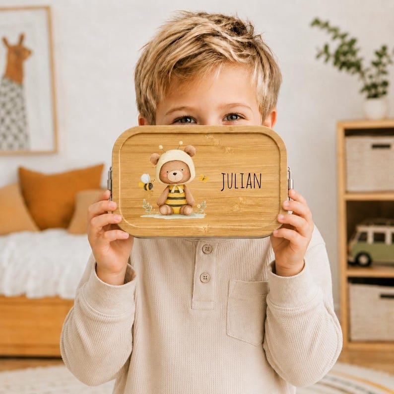 May include: A rectangular bamboo lunch box featuring a printed illustration of a teddy bear dressed in a bee costume with a small bee flying nearby. The name JULIAN is printed in black text on the right side of the light brown wooden lid.