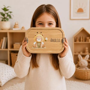 May include: A rectangular bamboo lunch box featuring a printed illustration of a bear wearing a bee costume and wings, accompanied by a small bee graphic. The name AMALIA is printed in black sans-serif text on the right side of the lid.