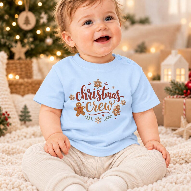 May include: A light blue short-sleeved t-shirt featuring the text Christmas Crew in brown script. The graphic includes illustrations of a gingerbread man, a star-shaped cookie, a tree-shaped cookie, and small snowflakes in brown and tan.