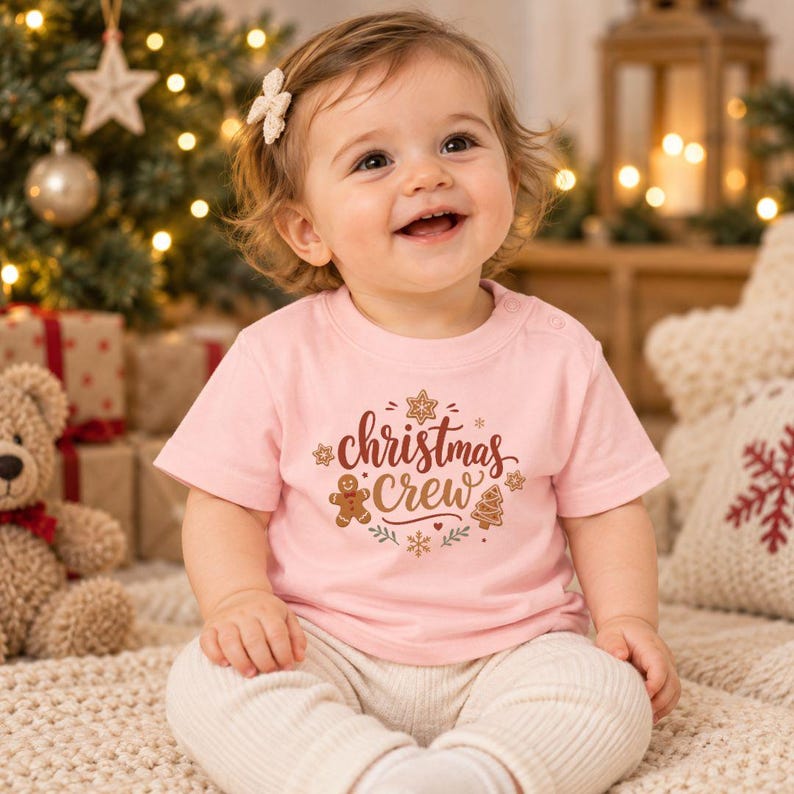 May include: A light pink short-sleeved t-shirt featuring the text "christmas crew" in dark red script, surrounded by illustrations of a gingerbread man, a gingerbread tree, and snowflakes in brown, red, and white colors.