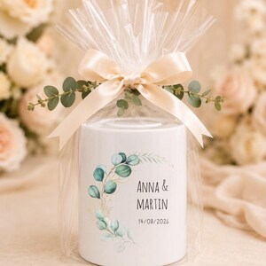 May include: A white cylindrical ceramic money box wrapped in clear plastic with a cream-coloured satin ribbon and eucalyptus sprigs. The front features a green eucalyptus leaf wreath design with the text ANNA & MARTIN and 14/08/2026.