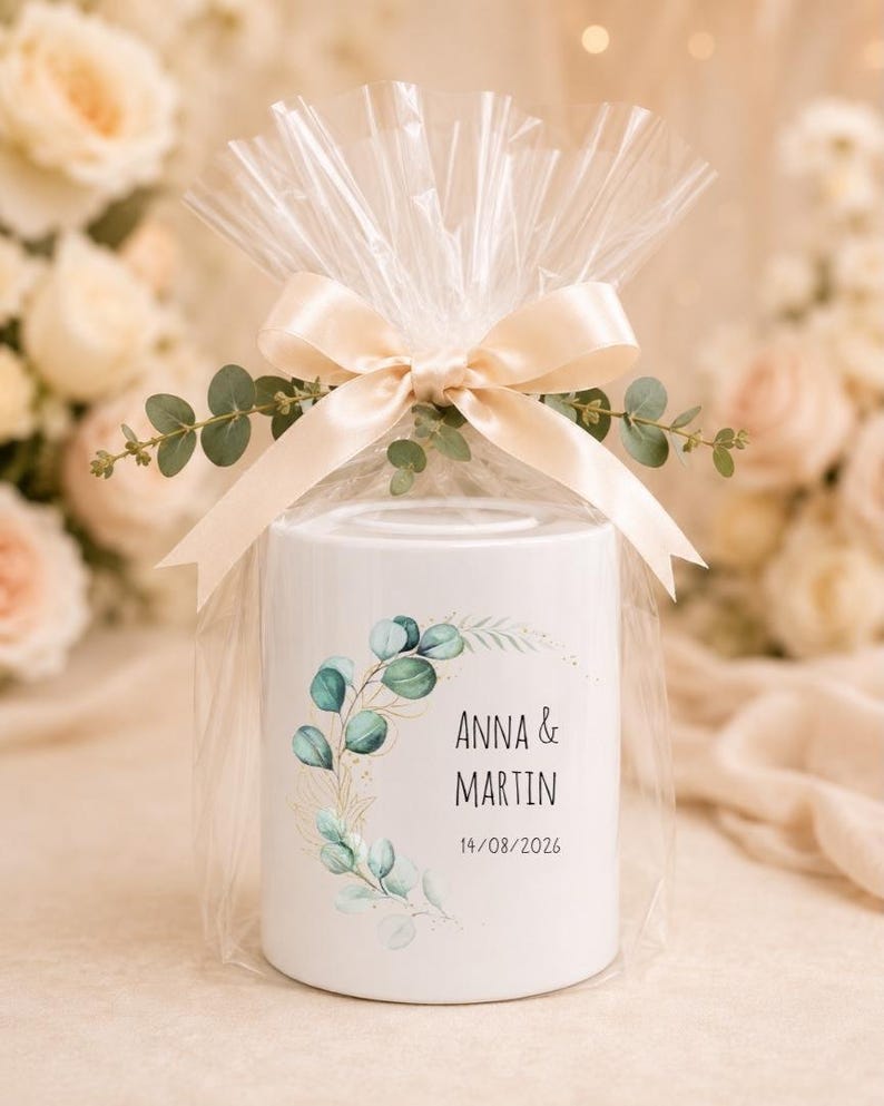 May include: A white cylindrical ceramic money box wrapped in clear plastic with a cream-coloured satin ribbon and eucalyptus sprigs. The front features a green eucalyptus leaf wreath design with the text ANNA & MARTIN and 14/08/2026.