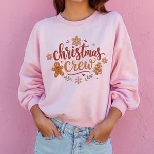 May include: A light pink crewneck sweatshirt featuring the text "christmas crew" in brown script. The design includes a gingerbread man, a gingerbread tree, snowflakes, and star-shaped cookies in brown, red, and green colors.
