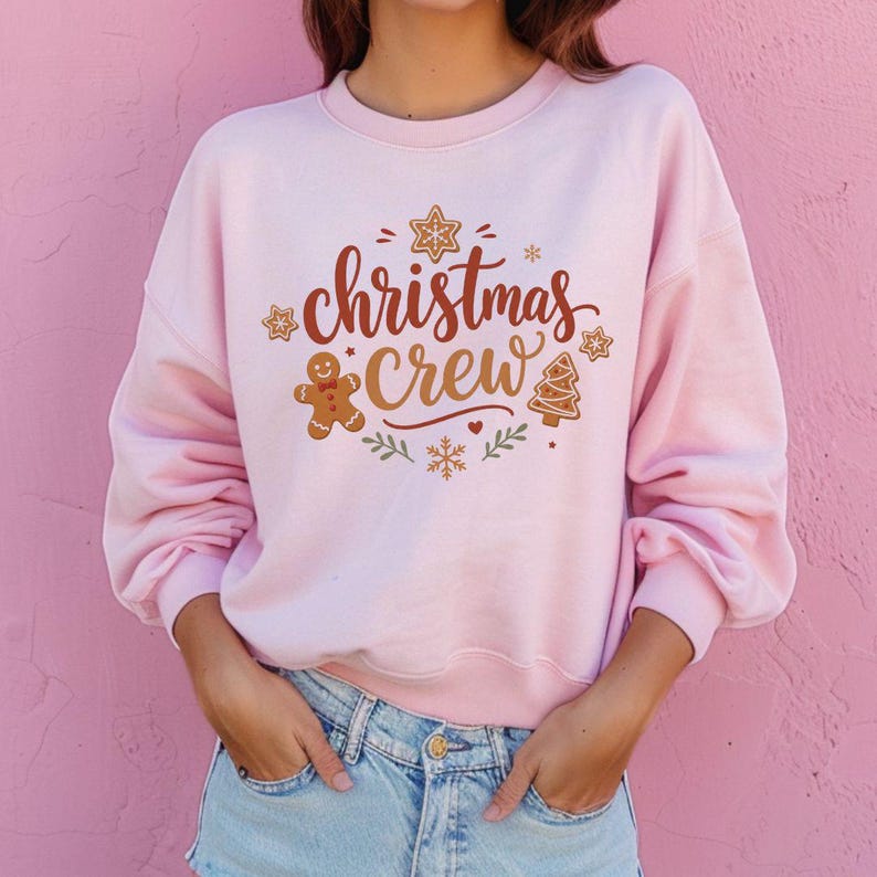 May include: A light pink crewneck sweatshirt featuring the text "christmas crew" in brown script. The design includes a gingerbread man, a gingerbread tree, snowflakes, and star-shaped cookies in brown, red, and green colors.