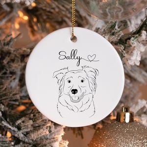May include: A white ceramic Christmas ornament with a black line drawing of a dog's face. The name "Sally" is written above the dog, with a heart symbol. The ornament hangs from a gold string.