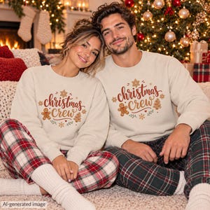 May include: Two beige crewneck sweatshirts featuring the text "christmas crew" in a brown script font, surrounded by graphic illustrations of a gingerbread man, a Christmas tree, a star, and snowflakes in brown, red, and green colors.