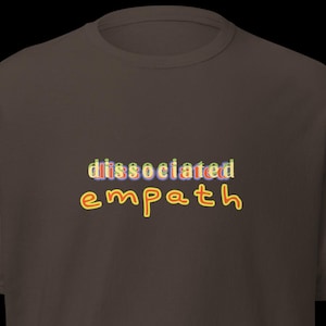 Könnte beinhalten: Ein braunes T-Shirt mit den Worten "dissociated empath" in bunten Buchstaben im Blasenstil. Der Text ist mittig auf der Vorderseite des Shirts platziert. Das Shirt hat kurze Ärmel und einen Rundhalsausschnitt.
