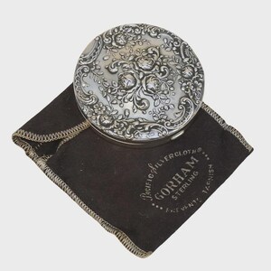 May include: A round, silver compact with an ornate floral design on the lid rests on a dark brown cloth. The cloth has a stitched border and the text "Pacific Silvercloth Gorham Sterling" printed on it.