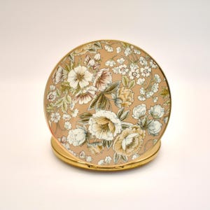 May include: A vintage compact mirror with a floral design. The round compact features a tan background with a pattern of white, cream, and yellow flowers with green leaves. The compact has a gold-colored metal frame.
