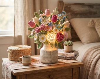 Könnte beinhalten: Ein gehäkelter Blumenstrauß in einer hellen Keramikvase mit integriertem Licht. Das Arrangement besteht aus gehäkelten Tulpen, Rosen und Gänseblümchen in Rosa, Gelb, Weiß und Blau mit grünen Blättern. Die Vase hat eine strukturierte Oberfläche.