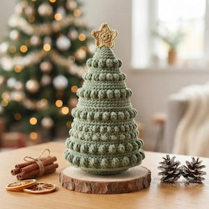 Op de afbeelding: Een saliegroene gehaakte kerstboom met een textuur van nopjessteken, bekroond met een klein gouden gehaakt sterretje. De boom is bevestigd op een ronde houten schijf als voet, wat een decoratief object vormt voor de feestdagen.