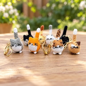 May include: A collection of eight small, round, crocheted cat keychains in various colors including gray, orange, white, brown, and black. Each cat features a white belly, black bead eyes, embroidered whiskers, and a vertical tail attached to a gold ring.