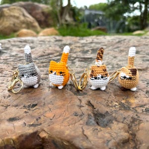 Puede incluir: Cuatro llaveros de gato hechos a mano en crochet en gris, naranja, marrón y beige. Cada gato tiene una cara y patas blancas, con un llavero dorado. Los gatos están sobre una superficie texturizada.