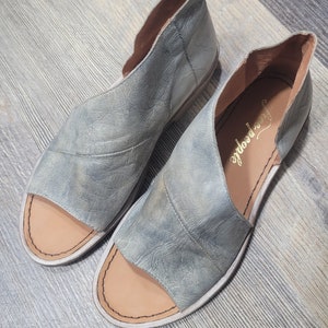K&ouml;nnte beinhalten: Ein Paar offene Sandalen aus abgenutztem, hellblauem Leder. Die Sandalen haben ein Ausschnittdesign an den Seiten und eine beige Innensohle. Der Markenname "free people" ist auf der Innensohle sichtbar. Die Sandalen werden auf einer Holzoberfl&auml;che pr&auml;sentiert.
