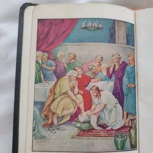 May include: Open book with a colorful illustration depicting a biblical scene. A man in a red robe washes the feet of another man, surrounded by other figures. Text at the bottom reads, "Jesus knowing...began to wash the disciples' feet..."