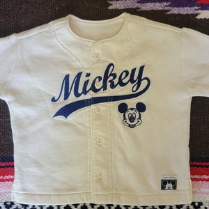 May include: Cream-colored baseball-style shirt with the name "Mickey" in navy blue script across the chest. A navy blue Mickey Mouse graphic is on the right side. The shirt has a button-up front and short sleeves. A small tag with a Mickey Mouse logo is on the bottom.