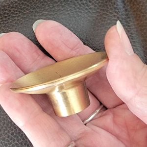 May include: A gold-toned metal cabinet knob featuring a wide, shallow, circular concave top surface that tapers down into a cylindrical base. The hardware has a smooth, polished finish and a simple, functional design for furniture or cabinetry.