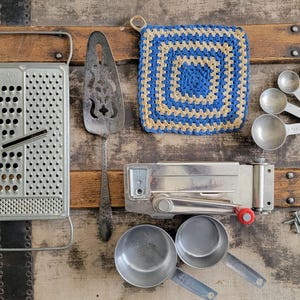 May include: An assortment of vintage kitchen tools, including a grater, pie server, crocheted pot holder, measuring spoons, a can opener, and measuring cups. The items are arranged on a wooden surface, showcasing a rustic aesthetic.