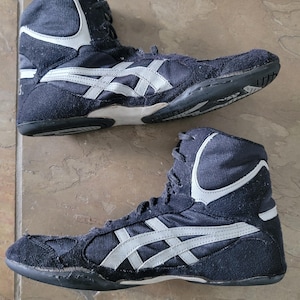 May include: Pair of navy blue wrestling shoes with white and silver accents. The high-top shoes feature a suede and mesh design, with the brand logo on the side. The soles are black and the shoes are designed for athletic performance.