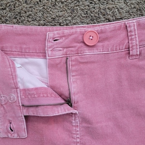 Op de afbeelding: Close-up van een roze corduroy rok met een knoopsluiting en een rits. De rok heeft een knoop op de tailleband en een kleine zak. De stof heeft een geribbelde textuur.