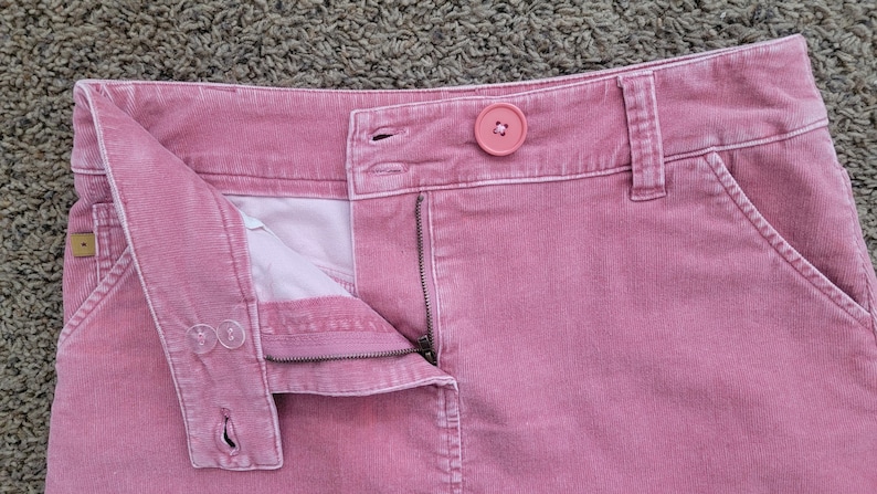 Op de afbeelding: Close-up van een roze corduroy rok met een knoopsluiting en een rits. De rok heeft een knoop op de tailleband en een kleine zak. De stof heeft een geribbelde textuur.