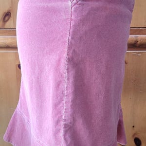 Op de afbeelding: Een stoffen rok in stoffig roze met een uitlopende zoom. De rok heeft een getailleerde taille, een voorzak met een klein goudkleurig sterdetail en een subtiele zoom. De rok is gemaakt van een geribbelde corduroy stof.