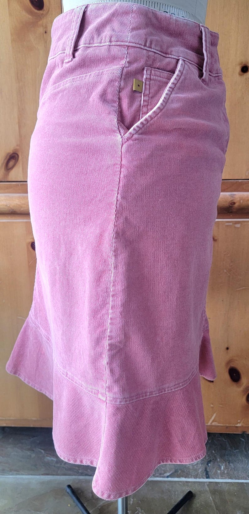 Op de afbeelding: Een stoffen rok in stoffig roze met een uitlopende zoom. De rok heeft een getailleerde taille, een voorzak met een klein goudkleurig sterdetail en een subtiele zoom. De rok is gemaakt van een geribbelde corduroy stof.