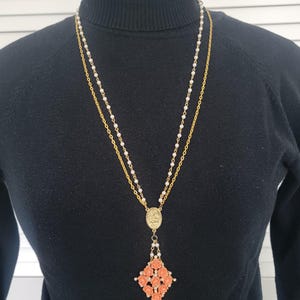 May include: A gold-tone necklace with a double chain design. The necklace features a pendant with a coral-colored floral arrangement and a small oval charm. The necklace is worn over a black turtleneck.