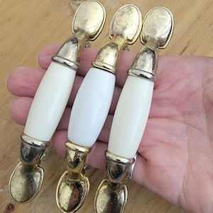 May include: Three cabinet pulls featuring a central white cylindrical section made of a smooth material, flanked by metallic gold-toned ends with a weathered finish. Each handle has a rounded mounting plate at both the top and the bottom.