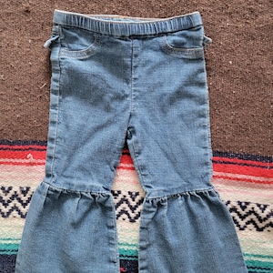 May include: Light blue denim pants with a flared leg design. The pants have an elastic waistband, two front pockets, and decorative stitching. The pants are designed for children.