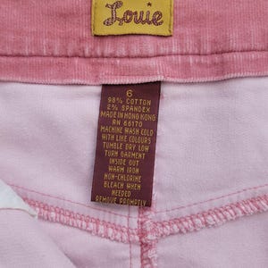 Op de afbeelding: Close-up van roze corduroy stof met een label met de tekst "Louie". Het rechthoekige label toont de merknaam in cursief, evenals onderhoudsinstructies en de samenstelling van de stof.