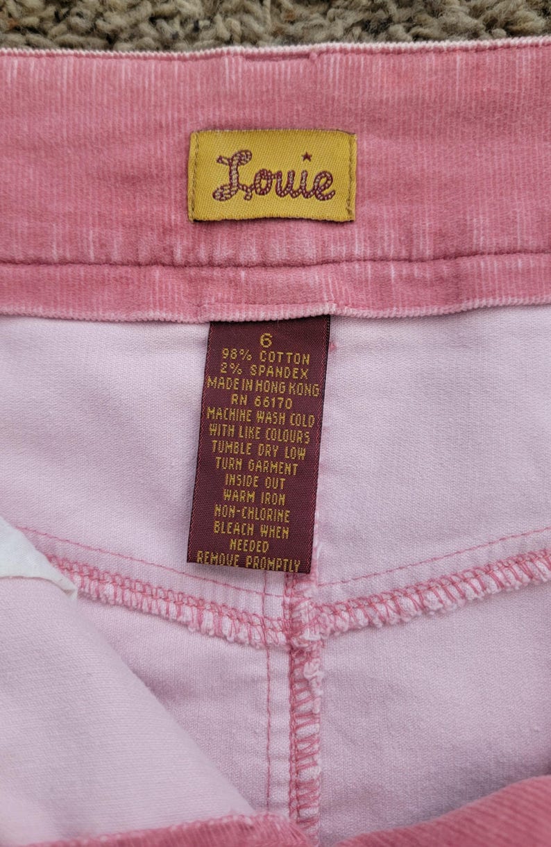 Op de afbeelding: Close-up van roze corduroy stof met een label met de tekst "Louie". Het rechthoekige label toont de merknaam in cursief, evenals onderhoudsinstructies en de samenstelling van de stof.