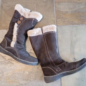 May include: A pair of dark brown mid-calf boots featuring a soft beige faux fur trim at the top and along the side. The boots include two round brown buttons on the upper shaft, visible side zippers, and white contrast stitching along the soles.