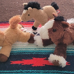 May include: Three plush toy horses in shades of brown, tan, and white are arranged on a patterned blanket. Each horse has a soft, furry texture, with details like black manes and tails. The toys are designed to resemble miniature horses.