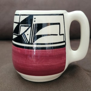 May include: A ceramic mug with a white background, a burgundy base, and a black and white geometric design. The mug has a large, rounded handle and is suitable for hot or cold beverages.