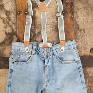 May include: Light blue denim shorts with attached green and white striped suspenders. The suspenders have brown leather accents and metal hardware. The shorts have a button closure and front pockets. The shorts are approximately 10 inches long.