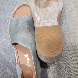 K&ouml;nnte beinhalten: Ein Paar offene Sandalen in einem ged&auml;mpften Blau-Grau-Ton. Die Sandalen haben ein Obermaterial aus Leder mit einem Ausschnitt-Design und eine braune Sohle. Der Markenname "free people" ist auf der Innensohle sichtbar. Die Sohle misst ca. 2.5 cm.