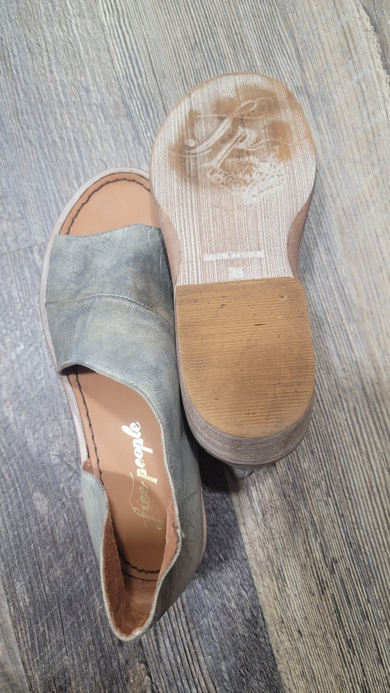 K&ouml;nnte beinhalten: Ein Paar offene Sandalen in einem ged&auml;mpften Blau-Grau-Ton. Die Sandalen haben ein Obermaterial aus Leder mit einem Ausschnitt-Design und eine braune Sohle. Der Markenname "free people" ist auf der Innensohle sichtbar. Die Sohle misst ca. 2.5 cm.