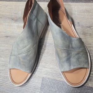K&ouml;nnte beinhalten: Ein Paar offene Sandalen aus abgenutztem, hellblauem Leder. Die Sandalen haben ein Cutout-Design und eine beige Innensohle. Der Markenname "Free People" ist auf der Innensohle einer Sandale sichtbar. Die Schuhe werden auf einer Holzoberfl&auml;che pr&auml;sentiert.