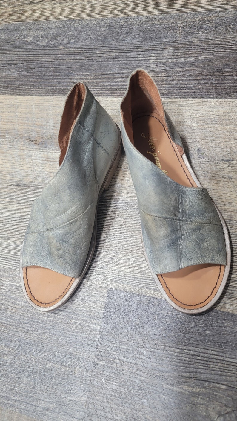 K&ouml;nnte beinhalten: Ein Paar offene Sandalen aus abgenutztem, hellblauem Leder. Die Sandalen haben ein Cutout-Design und eine beige Innensohle. Der Markenname "Free People" ist auf der Innensohle einer Sandale sichtbar. Die Schuhe werden auf einer Holzoberfl&auml;che pr&auml;sentiert.