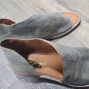 K&ouml;nnte beinhalten: Ein Paar offene Sandalen in einem ged&auml;mpften Gr&uuml;nton. Die Sandalen haben ein Cutout-Design und den Markennamen "Free People" in goldfarbenen Buchstaben. Die Sohlen sind hellbraun.