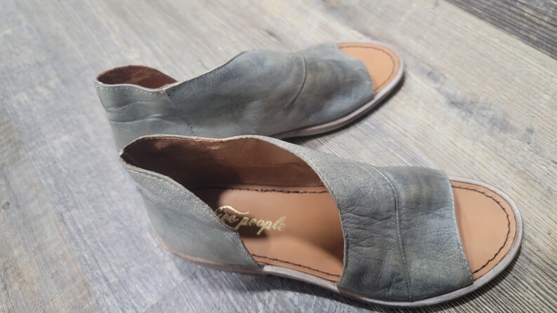K&ouml;nnte beinhalten: Ein Paar offene Sandalen in einem ged&auml;mpften Gr&uuml;nton. Die Sandalen haben ein Cutout-Design und den Markennamen "Free People" in goldfarbenen Buchstaben. Die Sohlen sind hellbraun.