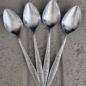 May include: Four silver-toned metal spoons arranged in a fan shape. Each spoon features a polished, smooth oval bowl and a slender handle decorated with an embossed floral pattern consisting of small leaves and blossoms along the entire length.