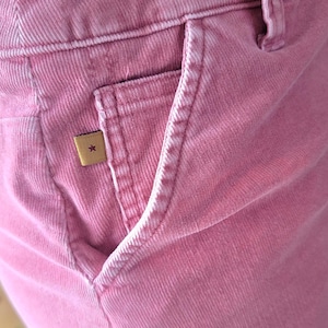 Op de afbeelding: Close-up van een roze corduroy broek met een voorzak. De broek heeft een klein, vierkant label met een sterembleem. De stof heeft een geribbelde textuur. De broek heeft een stoffige roze kleur.