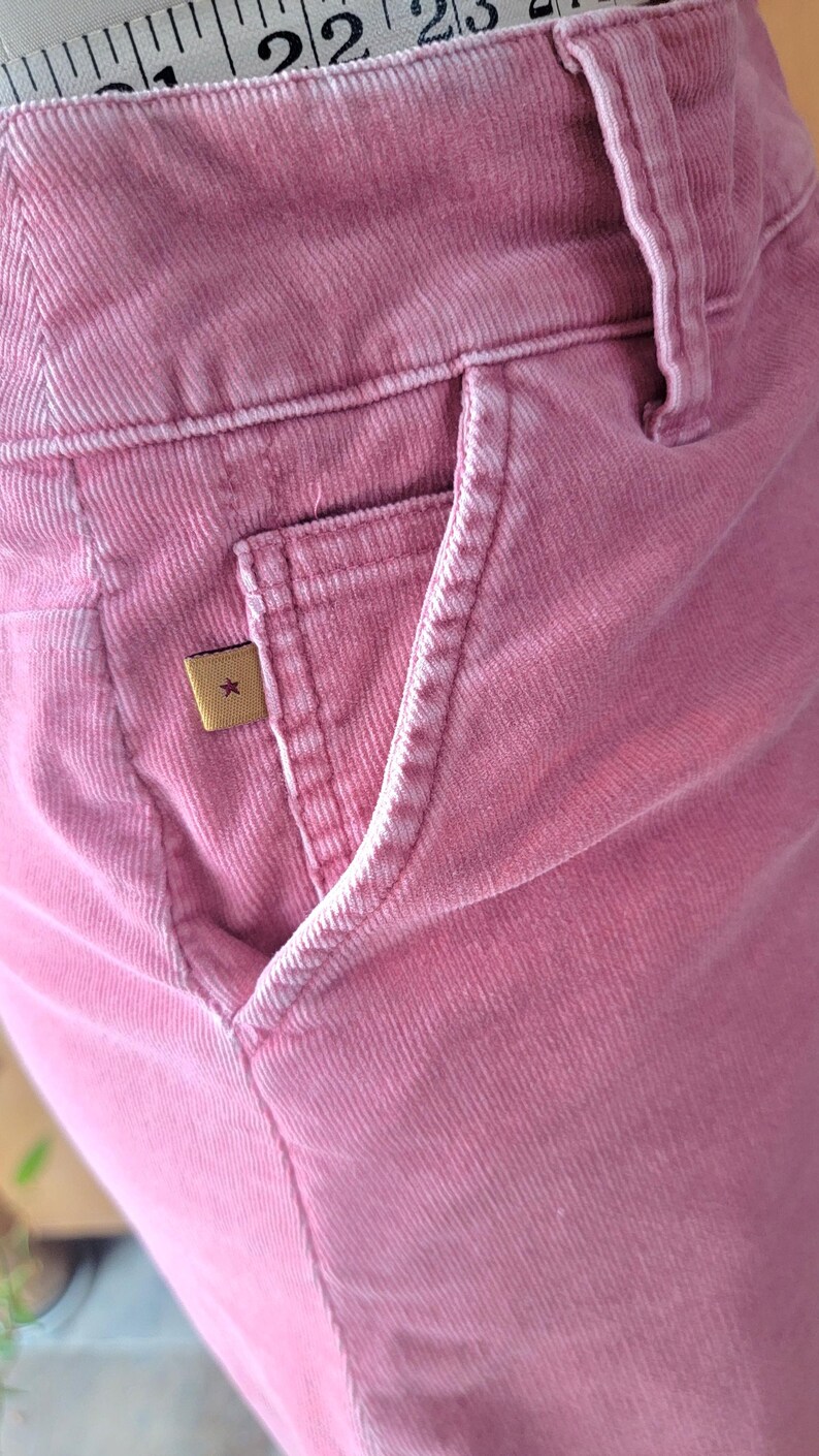 Op de afbeelding: Close-up van een roze corduroy broek met een voorzak. De broek heeft een klein, vierkant label met een sterembleem. De stof heeft een geribbelde textuur. De broek heeft een stoffige roze kleur.