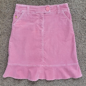 May include: A pink corduroy skirt with a button closure and pockets. The skirt has a slight A-line shape and a ruffled hem. The skirt is made of a soft, textured fabric.